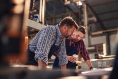 Twee mannen bespreken samen werkdocumenten aan een tafel in een industri&euml;le werkplaats.