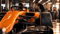 Oranje racewagen in een werkplaats, met een monteur die op de achtergrond aan de wagen werkt.